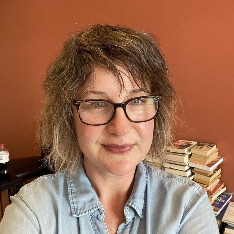 Person smiling in office with stacked books.